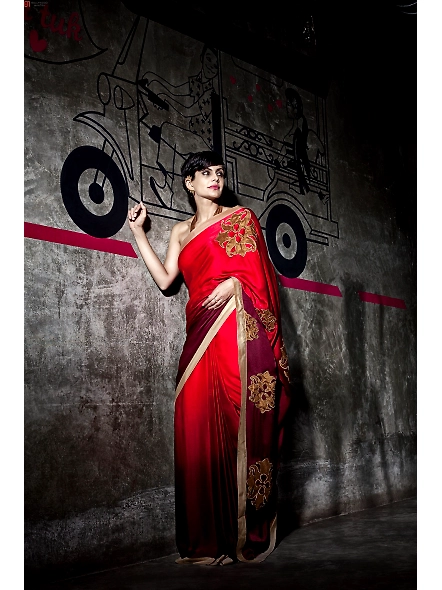 A woman in a red Georgette and silk sari stands beside a wall, showcasing her elegant attire and raw