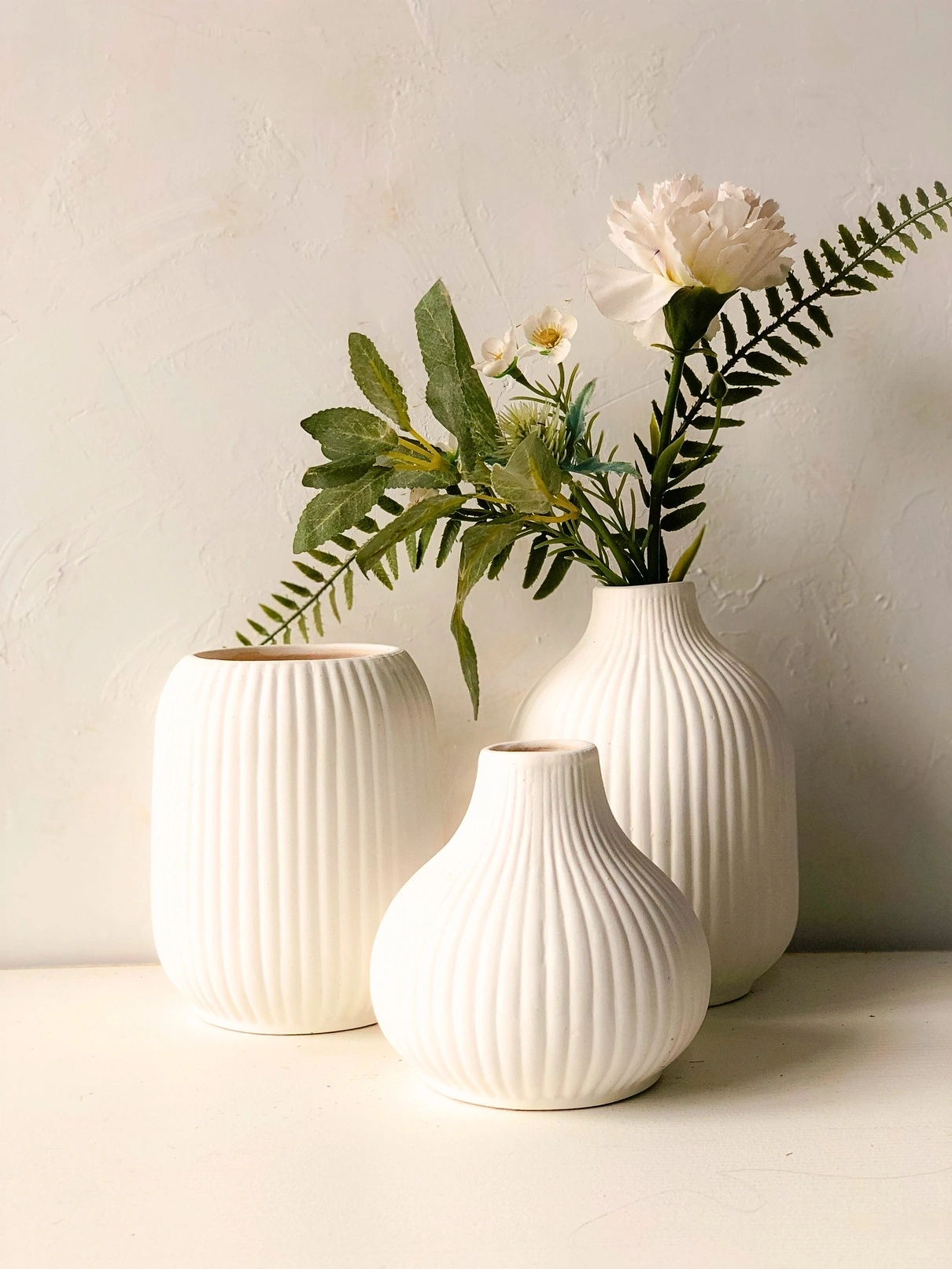 Three white, ribbed vases with flowers and greenery against a textured wall.