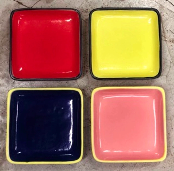 Four colorful square dishes in red, yellow, blue, and pink on a concrete surface.