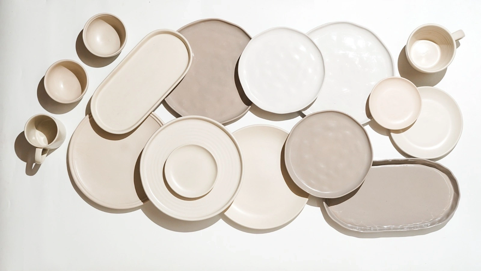 Assorted ceramic dishes and cups in neutral tones arranged on a white background.