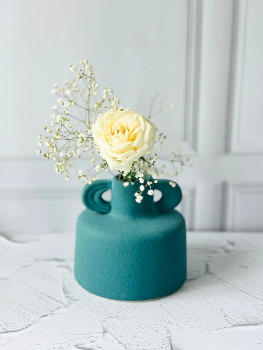 A single yellow rose and baby's breath in a teal vase on a white surface.