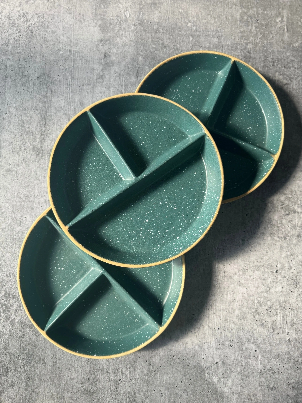 Three teal, sectioned ceramic plates with a gold rim, stacked on a concrete surface.