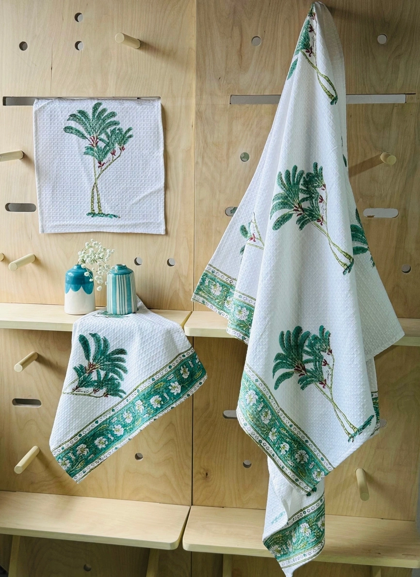 A wooden pegboard with embroidered palm tree textiles: a hanging towel, a folded cloth, and vases on