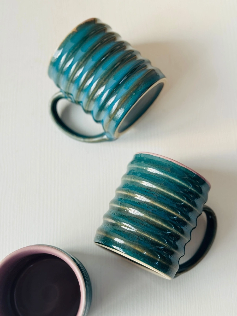 Three ceramic mugs with blue glazing, two standing and one toppled, on a white surface.