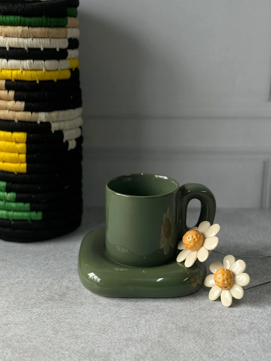 A green ceramic cup with saucer and two decorative flowers on a tabletop, with a woven basket in the