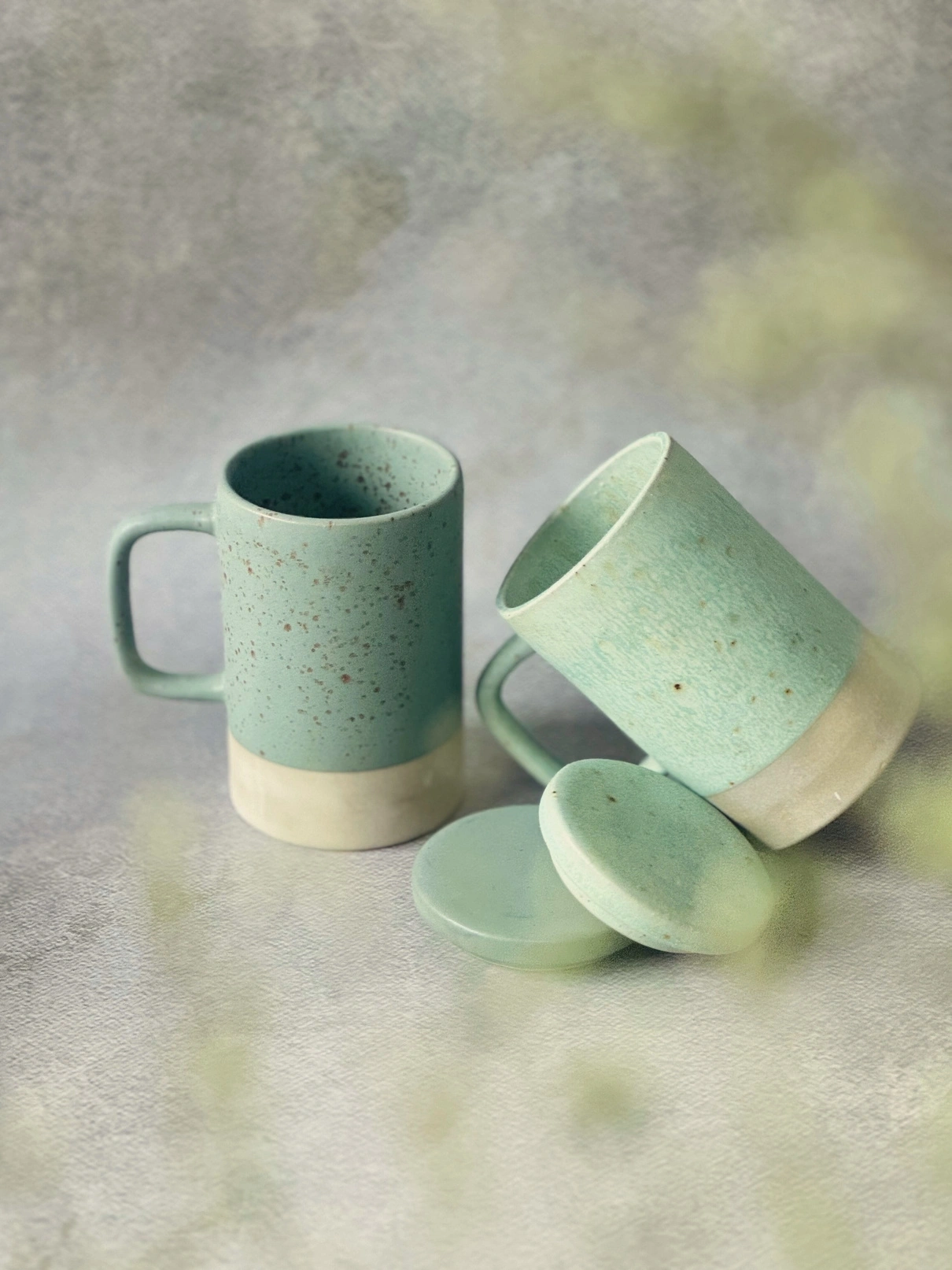 Two speckled mint green ceramic mugs with coasters on a textured background.