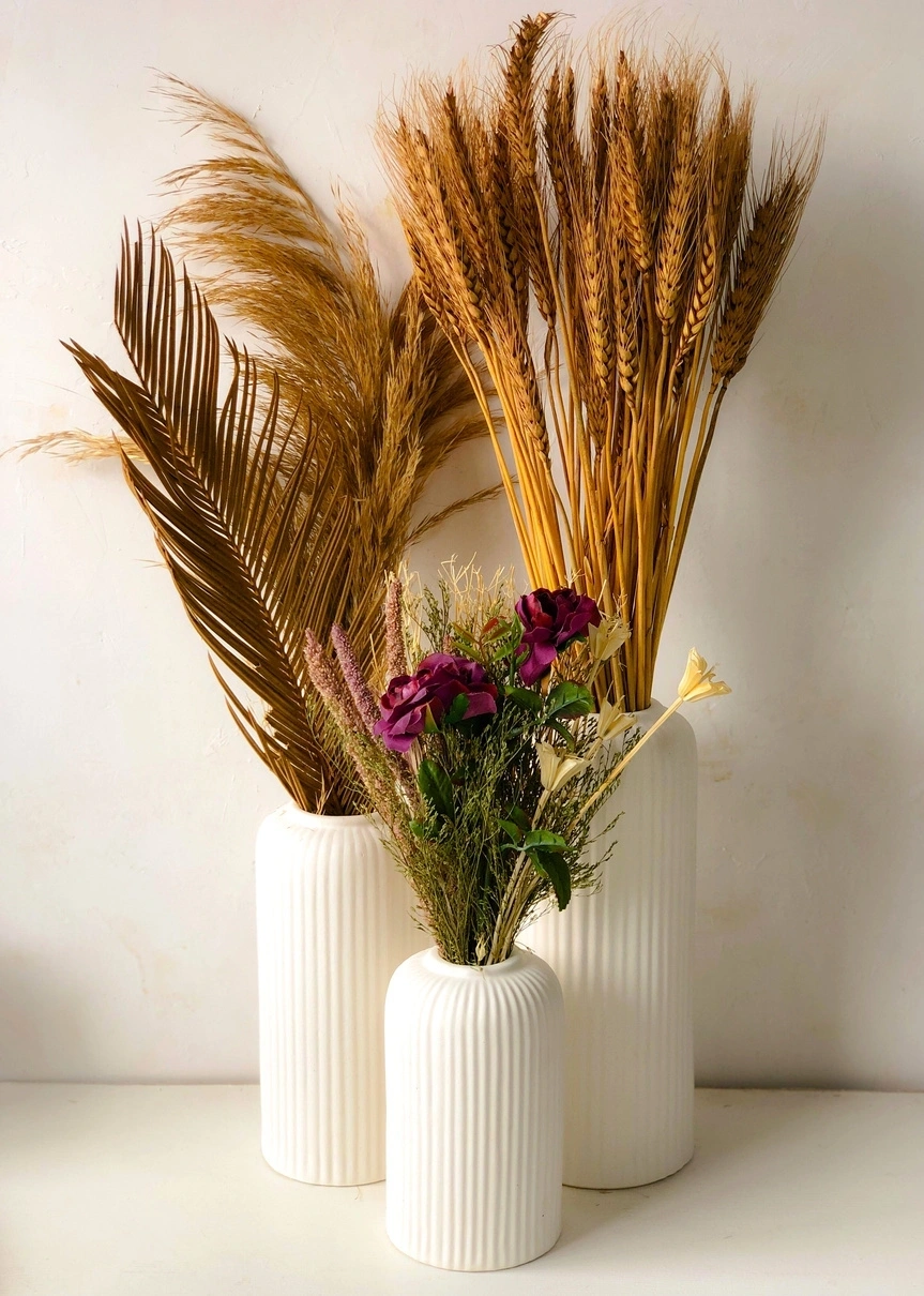 Three white ribbed vases holding wheat, pampas grass, and flowers against a cream backdrop.