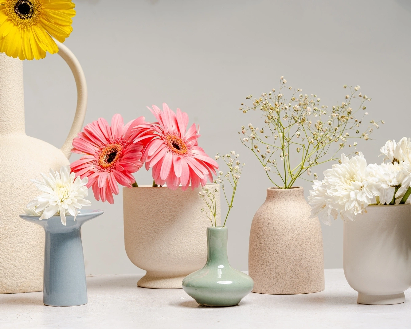 Assorted flowers in ceramic vases of different shapes and colors on a light background.