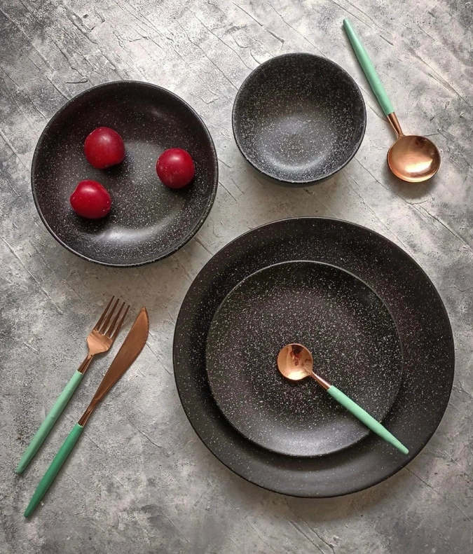 A table setting with black speckled dishes, red plums, and pastel green-handled cutlery.