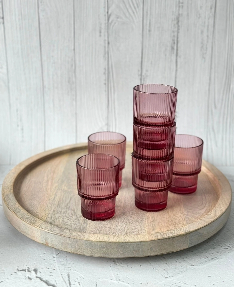 Six pink ribbed glasses on a round wooden tray against a white wooden backdrop.