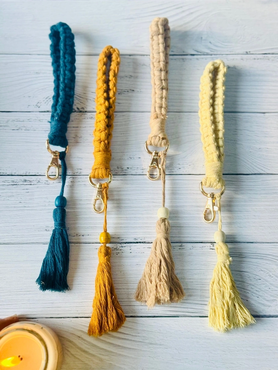 Four braided keychains with tassels, in blue, mustard, and cream colors, on a wooden background.