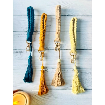 Four braided keychains with tassels, in blue, mustard, and cream colors, on a wooden background.