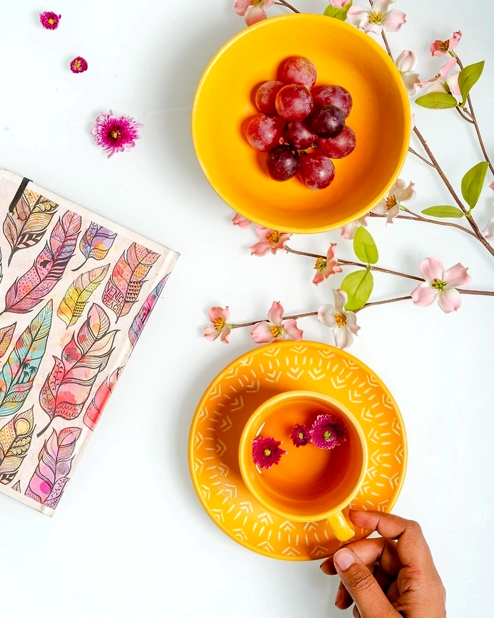 Yellow cup with flowers on saucer, grapes in bowl, floral branches, and patterned napkin on white ba