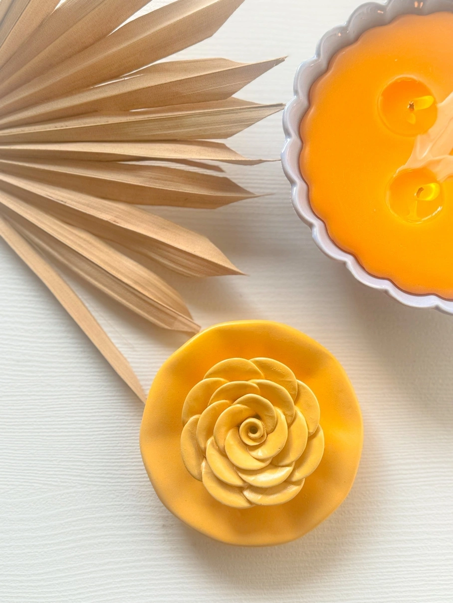 A decorative yellow candle with a rose design next to a lit scalloped candle and dried palm leaves.