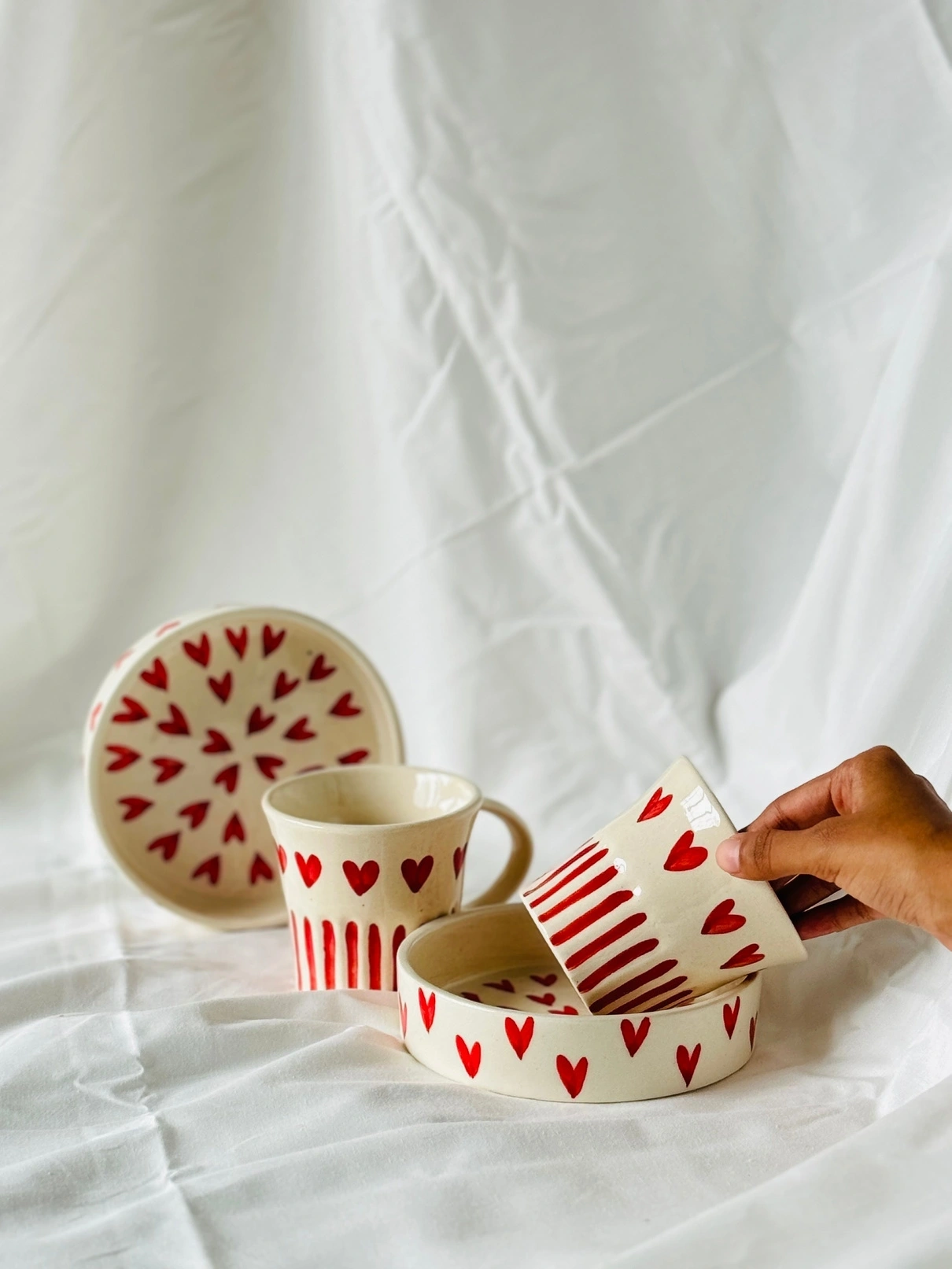 A hand tilting a heart-patterned mug with matching saucers on a white fabric.