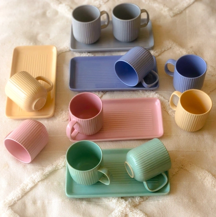 Assorted pastel-colored mugs and trays with ribbed designs on a textured cloth.